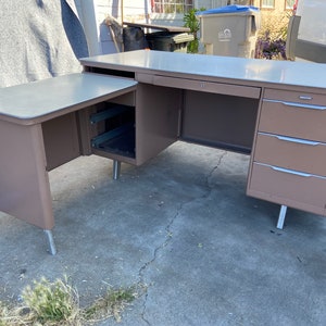 May include: A vintage L-shaped desk with a light brown finish and a light gray top. The desk features a pull-out drawer and three additional drawers on the right side. The desk is supported by silver metal legs. A classic piece for a home office.