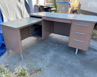 May include: A vintage L-shaped desk with a light brown finish and a light gray top. The desk features a pull-out drawer and three additional drawers on the right side. The desk is supported by silver metal legs. A classic piece for a home office.