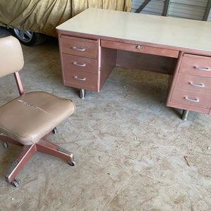 May include: A vintage metal desk with a light brown finish and a white laminate top. The desk has three drawers on each side and a single drawer in the center. A matching light brown swivel chair with a padded seat is in the foreground.