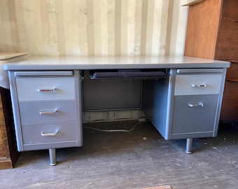 May include: A vintage metal desk with a light gray top and a dark gray frame. The desk has three drawers on the left and two drawers on the right, all with silver handles. The desk has a pull-out keyboard tray.