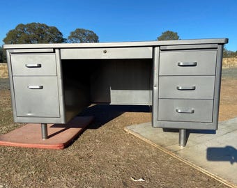 May include: A vintage metal desk with a silver finish. The desk has two sets of drawers on either side and a central drawer with a keyhole. The desk is supported by silver legs and sits on a red mat and a wooden board.