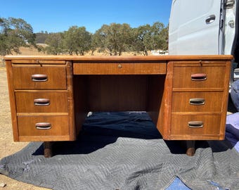May include: A vintage wooden desk with a warm brown finish. It features multiple drawers on each side and a central drawer with a keyhole. The desk is supported by short, rounded legs, and is set outdoors.
