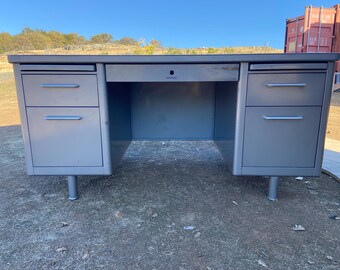 May include: A gray metal desk with two sets of drawers and a central drawer. The desk has a rectangular top and is supported by cylindrical legs. The desk is outdoors with a clear blue sky in the background.