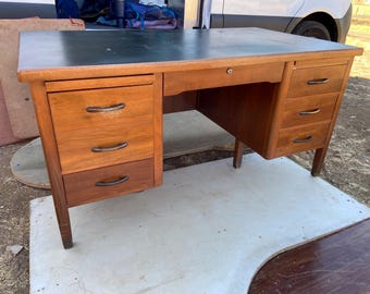 May include: A vintage wooden desk with a dark green top and multiple drawers. The desk has a warm, reddish-brown finish and metal drawer pulls. It features a keyhole and is supported by four legs.