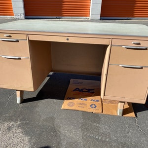 May include: A vintage metal desk with a light brown finish. The desk has a large work surface, three drawers on the left side, and two drawers on the right side. The desk has a metal frame and legs.