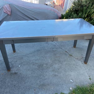 May include: A rectangular gray table with four legs. The table has a light gray surface and a darker gray frame. The legs are straight and end with brown feet. The table is outdoors on a concrete surface.