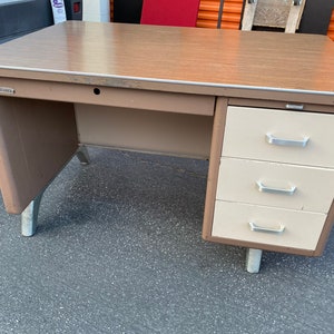 May include: A brown metal desk with three drawers and silver handles. The desk has a wood-grain laminate top and silver legs.