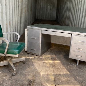 May include: A vintage metal desk with three drawers on the right side and one drawer on the left side. The desk has a light gray finish and is paired with a green swivel chair.