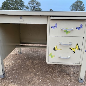 May include: A vintage metal desk with a light beige finish. The desk has three drawers with silver handles and butterfly decals. The top drawer has a keyhole.