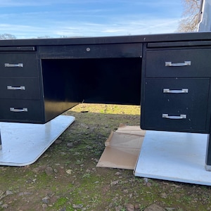 May include: A black metal desk with three drawers on each side and a central drawer. The desk has a rectangular shape and is supported by four legs. The drawers have silver handles. The desk is sitting on two white rectangular platforms.