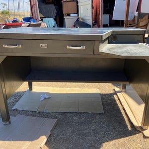 May include: A dark green metal desk with two drawers, a small side surface, and a lower shelf. The desk has a rectangular shape with a dark surface and silver drawer pulls. The desk is supported by four legs with gold-colored feet.
