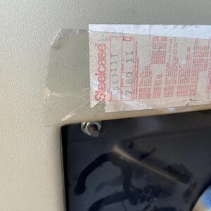 Puede incluir: Primer plano de una pegatina con la palabra "Steelcase" en rojo y blanco. La pegatina tiene varias l&iacute;neas de texto y n&uacute;meros. La pegatina est&aacute; adherida a una superficie beige claro con una superficie negra debajo.