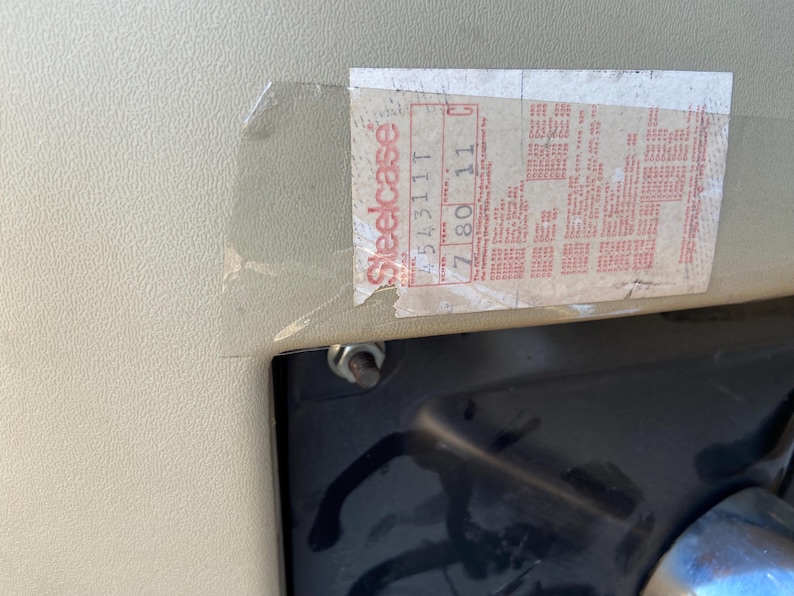 Puede incluir: Primer plano de una pegatina con la palabra "Steelcase" en rojo y blanco. La pegatina tiene varias l&iacute;neas de texto y n&uacute;meros. La pegatina est&aacute; adherida a una superficie beige claro con una superficie negra debajo.