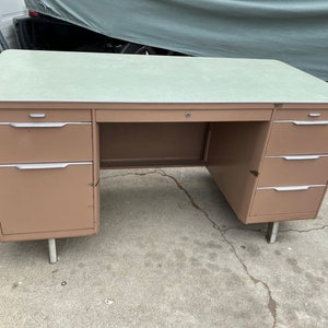 May include: A vintage metal desk with a light green top and a tan-colored body. The desk has multiple drawers with silver handles and rests on silver legs. The desk is a mid-century modern style.