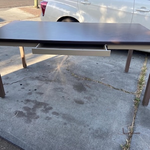 May include: A rectangular desk with a dark brown tabletop and a beige drawer. The legs are a contrasting brown colour. The desk is set on a concrete surface.