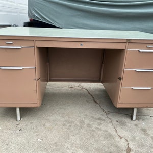 May include: A vintage metal desk with a light brown finish and a light green top. The desk has multiple drawers with silver handles and rests on silver legs. The desk has a key lock on the center drawer.