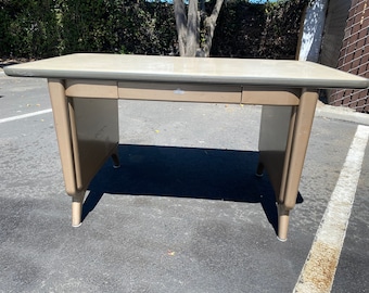 May include: A vintage metal desk in a neutral beige color. The desk has a rectangular top with a single drawer and rounded legs. The desk is approximately 48 inches wide and 24 inches deep.