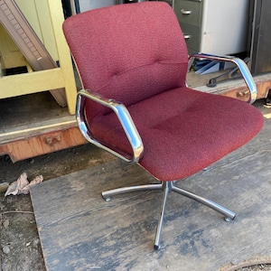 Puede incluir: Una silla de oficina burdeos con respaldo bajo y brazos y base cromados. La silla tiene un asiento y respaldo de tela texturizada con detalles de botones. La base tiene cinco patas.