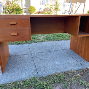 May include: A mid-century modern desk made of wood, featuring two drawers on the left side and an open shelf on the right. The desk has a warm, reddish-brown finish and is set on a concrete surface.