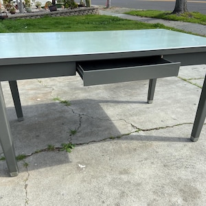 May include: A gray metal desk with a light gray top and a single drawer. The desk has a simple design and is in good condition.