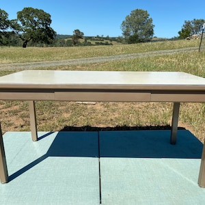 May include: A rectangular table with a light beige top and a brown frame. The table has a single drawer and is supported by four angled legs. The table is approximately 60 inches long and 30 inches wide.