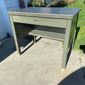 May include: A vintage olive green metal desk with a single drawer and a lower shelf. The desk has a rectangular top with a silver trim and is supported by four straight legs. The desk is outdoors on a concrete surface.