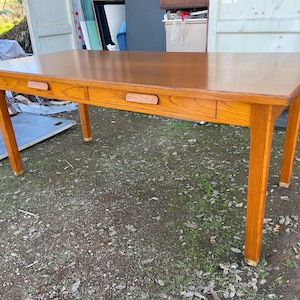 May include: A rectangular wooden table with a light brown finish. The table has two drawers with wooden handles and four angled legs. The table is set on a gravel surface.