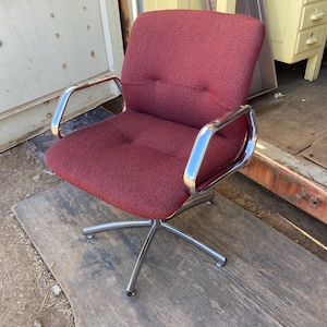 Puede incluir: Una silla de oficina burdeos con base de cinco estrellas y reposabrazos cromados. La silla tiene asiento y respaldo de tela texturizada. La silla est&aacute; sobre una superficie de madera.