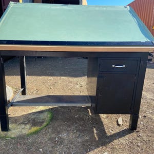 May include: A black drafting table with a green angled top. The table has a black metal frame, a drawer, and a cabinet. The top is supported by a wooden frame. The table is designed for drawing and drafting.