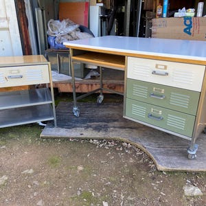 Pode incluir: Uma escrivaninha de metal branca e verde com três gavetas e uma tampa branca. A escrivaninha está sobre rodas e tem uma pequena prateleira de madeira por baixo. Há uma prateleira de metal menor com uma tampa de madeira e duas prateleiras atrás da escrivaninha.