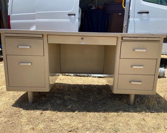 May include: A vintage metal desk in a light beige color. It features a central drawer with a keyhole and multiple drawers on each side. The desk has a silver-colored top and rests on short, cylindrical legs.