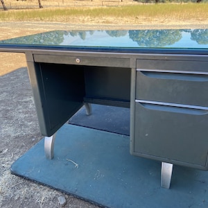 May include: A vintage metal desk with a glass top. The desk is painted a dark green color and has two drawers on the right side. The legs are silver and the desk has a key lock.
