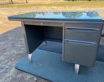 May include: A vintage metal desk with a glass top. The desk is painted a dark green color and has two drawers on the right side. The legs are silver and the desk has a key lock.