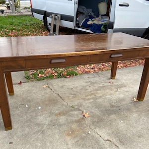 May include: A rectangular wooden desk with two drawers and four legs. The desk is a medium brown color with a glossy finish. The legs have gold-colored accents at the bottom. The desk is outdoors on a concrete surface.