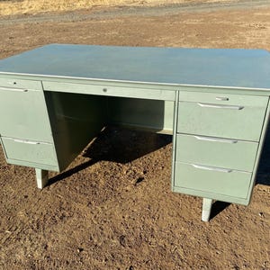 May include: A vintage metal desk with a light green finish. The desk has a large work surface and three drawers on the right side. The desk is on a brown dirt surface.