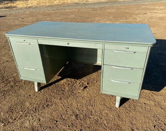 May include: A vintage metal desk with a light green finish. The desk has a large work surface and three drawers on the right side. The desk is on a brown dirt surface.