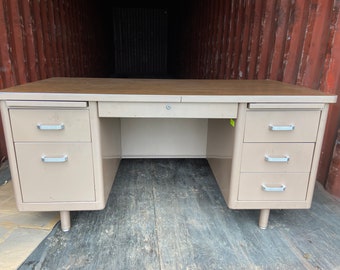 May include: A beige metal desk with a brown laminate top and three drawers on each side. The desk has a simple design and is in good condition.