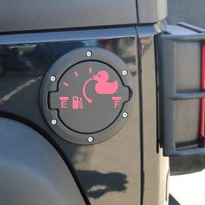 May include: A circular black fuel door cover for a vehicle featuring a pink decal of a fuel gauge. The gauge includes the letters E and F, a fuel pump icon, and a pink rubber duck graphic positioned at the full mark, connected by a curved line.