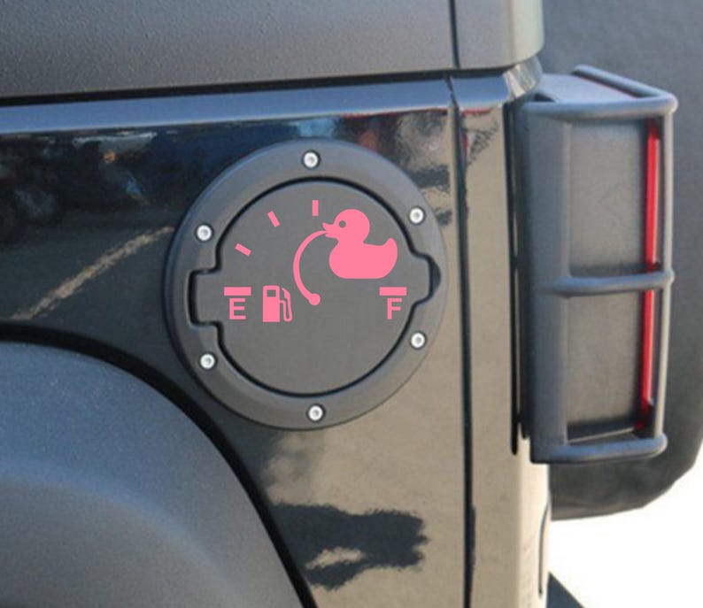 May include: A circular black fuel door cover for a vehicle featuring a pink decal of a fuel gauge. The gauge includes the letters E and F, a fuel pump icon, and a pink rubber duck graphic positioned at the full mark, connected by a curved line.