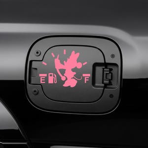 May include: A black fuel door with a pink Minnie Mouse graphic. The graphic replaces the fuel gauge and includes the letters "E" and "F" for empty and full, and a gas pump icon. The fuel door is on a black vehicle.
