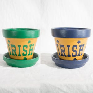May include: Two terracotta plant pots with matching saucers. One pot features a green rim and base with yellow sides, green shamrocks, and the word IRISH in green. The other features a blue rim and base with yellow sides, blue shamrocks, and IRISH in blue.