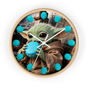 May include: A round wall clock with a light wood frame featuring a printed image of a green creature with large dark eyes eating a bright blue macaron. The clock face uses twelve small blue macarons as hour markers and has white clock hands.