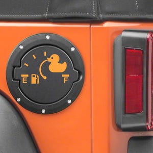 May include: A black circular fuel door cover for a vehicle, featuring an orange graphic of a rubber duck replacing the fuel gauge needle. The design includes the letters E and F, a fuel pump icon, and a curved line connecting to the duck.