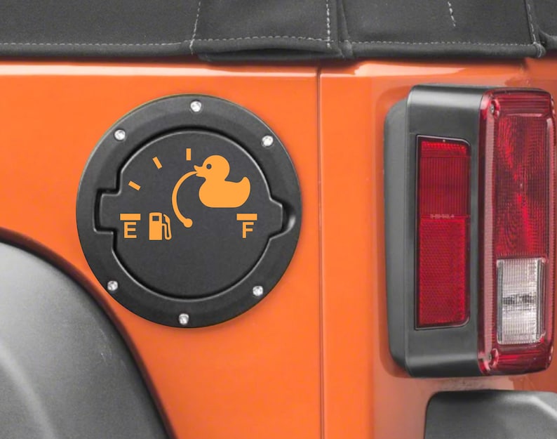 May include: A black circular fuel door cover for a vehicle, featuring an orange graphic of a rubber duck replacing the fuel gauge needle. The design includes the letters E and F, a fuel pump icon, and a curved line connecting to the duck.