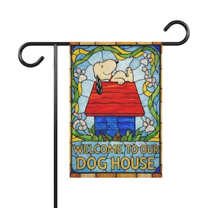 May include: A garden flag featuring Snoopy relaxing on his red doghouse, with the words "WELCOME TO OUR DOG HOUSE". The flag has a stained-glass style design with a black metal stand.