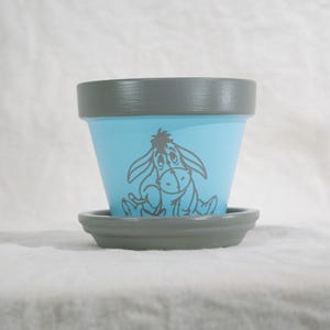 May include: A hand-painted ceramic flower pot with a light blue exterior and a gray rim and matching saucer. The pot features a gray illustration of Eeyore from Winnie the Pooh. A decorative item for home or garden.