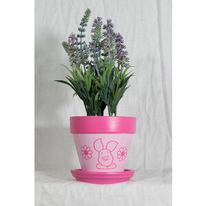 May include: Pink ceramic flower pot with a cartoon pig design and a saucer. The pot is filled with a green plant with purple flowers.