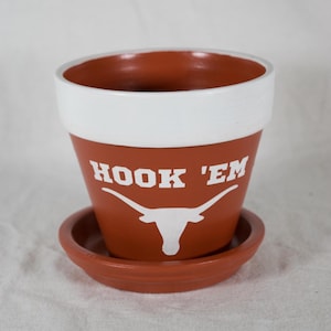 May include: A terracotta flower pot with a white rim and a white design of a longhorn steer with the words "Hook 'Em" on the side. The pot has a matching terracotta saucer.