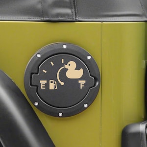 May include: A circular black fuel door cover for a vehicle, featuring a beige graphic of a rubber duck connected to a fuel pump icon. The design includes the letters E and F, mimicking a fuel gauge, with small silver rivets around the perimeter.