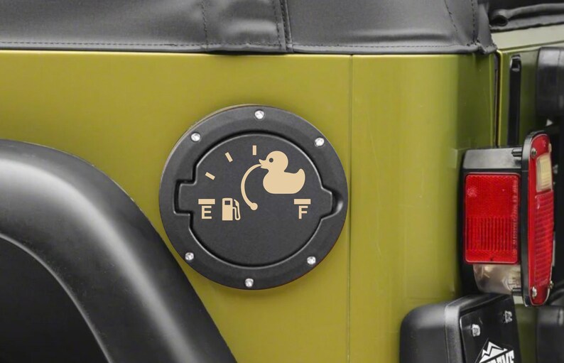 May include: A circular black fuel door cover for a vehicle, featuring a beige graphic of a rubber duck connected to a fuel pump icon. The design includes the letters E and F, mimicking a fuel gauge, with small silver rivets around the perimeter.